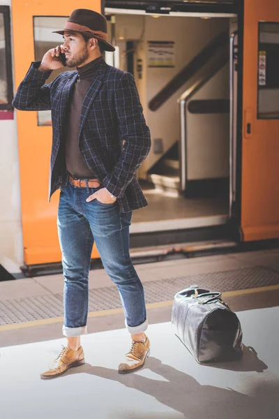 Atractivo y elegante joven hispano sosteniendo un teléfono inteligente en su mano y llamando mientras está de pie en la plataforma ferroviaria esperando el tren. . — Foto de Stock