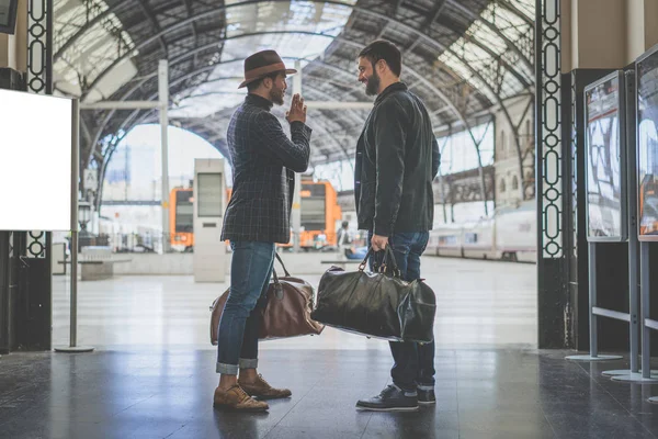 Dos empresarios barbudos multi-étnicos confiados juntos usando ropa casual y sosteniendo bolsas de viaje en las manos esperando el tren en la plataforma ferroviaria — Foto de Stock