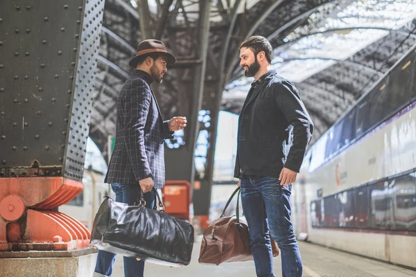 Dos hombres de negocios barbudos multi-étnicos sonrientes juntos usando ropa casual y sosteniendo bolsas de viaje en las manos esperando el tren en la plataforma ferroviaria — Foto de Stock