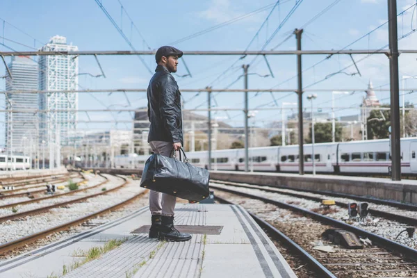 Atractivo hombre hispano confiado teniendo una mochila delante de él con una bolsa de ropa, caminando al lado de una plataforma en la estación de tren . — Foto de Stock