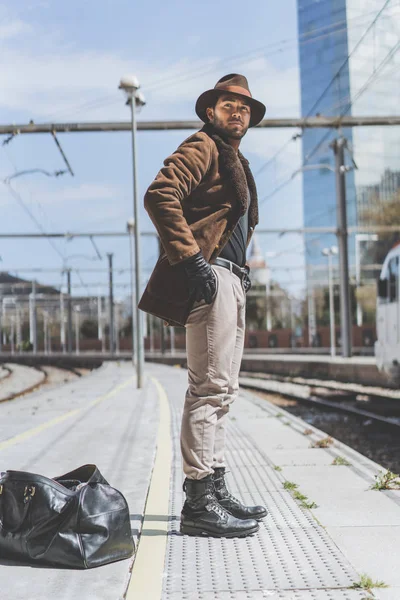 Atractivo hombre hispano confiado usando sombrero con una bolsa de ropa detrás de su espalda, esperando tren en la plataforma . — Foto de Stock