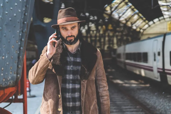 Atractivo hombre caucásico adulto barbudo sosteniendo el teléfono inteligente en su mano y llamando mientras está de pie en la plataforma ferroviaria esperando el tren — Foto de Stock