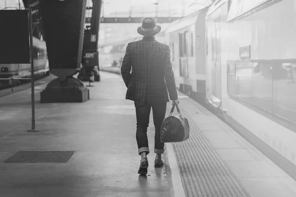 Joven hombre de negocios hispano confiado con bolsa de viaje con ropa casual y sombrero mirando el tren en la plataforma ferroviaria. Blanco y negro . — Foto de Stock
