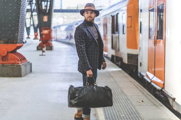 Atractivo empresario hispano confiado con bolsa de viaje con ropa casual y sombrero esperando el tren en la plataforma ferroviaria — Foto de Stock