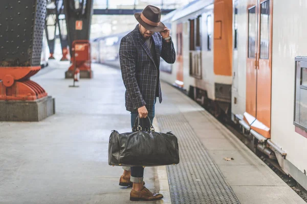 Atractivo hipster hispano confiado con bolsa de viaje con ropa casual y sombrero esperando el tren en la plataforma ferroviaria — Foto de Stock