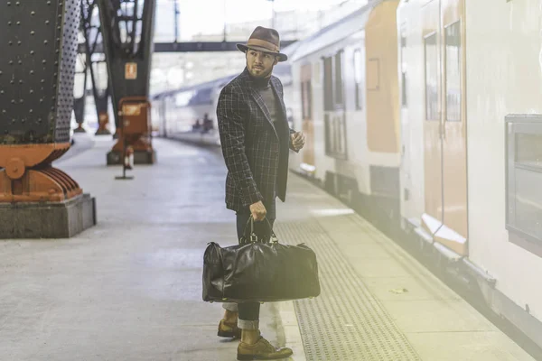 Guapo hipster hispano confiado con bolsa de viaje con ropa casual y sombrero esperando el tren en la plataforma ferroviaria — Foto de Stock
