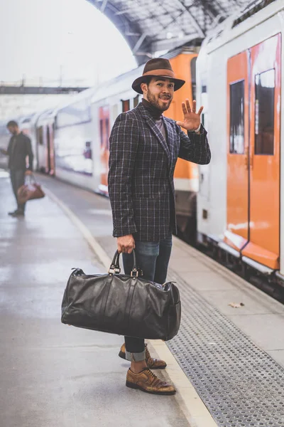Atractivo hipster hispano confiado con bolsa de viaje con ropa casual y sombrero dice adiós a los amigos en la plataforma ferroviaria — Foto de Stock