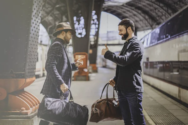 Dos felices hombres de negocios barbudos multiétnicos que llevan ropa casual y llevan bolsas de viaje en las manos. Amigos conversando casualmente mientras esperan el tren en la plataforma del tren — Foto de Stock