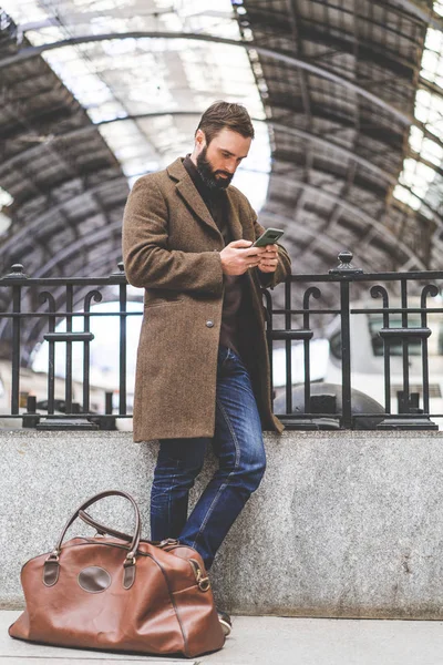 Hombre barbudo seguro de pie con bolsa de viaje en la sala de la estación de tren y mensaje de texto en su teléfono móvil . — Foto de Stock