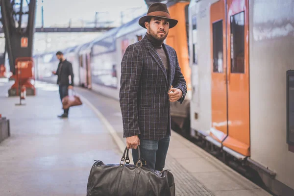 Atractivo hombre de negocios hispano confiado con bolsa de viaje con ropa casual y sombrero esperando el tren en la plataforma ferroviaria — Foto de Stock
