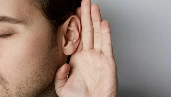 Слушающий самец держит руку возле уха на сером фоне. . — стоковое фото