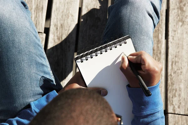 Atraente casualmente vestido jovem estudante negro do sexo masculino fazendo anotações em copybook, preparando-se para a lição na universidade . — Fotografia de Stock