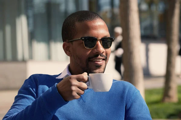 Retrato de un joven empresario afro-americano sonriente y confiado en sí mismo con ropa formal sentado en el banco en el parque de la ciudad y bebiendo café caliente. Cultivado . —  Fotos de Stock