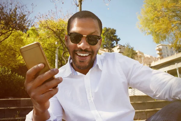 Junger fröhlich lächelnder afrikanisch-amerikanischer Mann in Sonnenbrille mit Handy-Smartphone im Stadtgarten. — Stockfoto