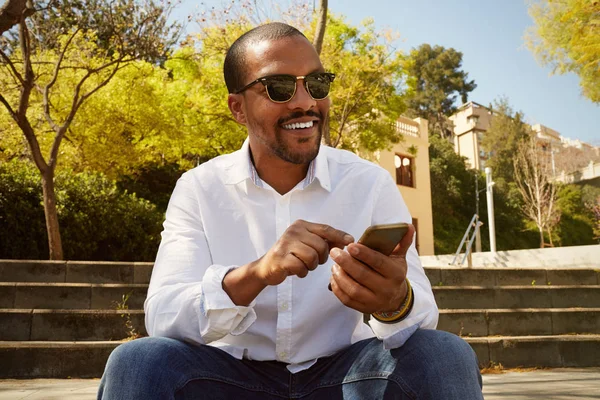 Junger selbstbewusster afrikanischer Mann zeigt auf Smartphone, während er im sonnigen Stadtpark sitzt. Konzept glücklicher Geschäftsleute, die draußen arbeiten — Stockfoto