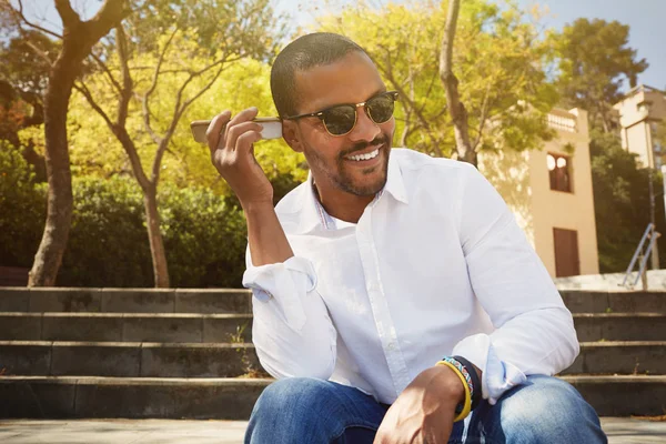 Feliz joven africano con camisa blanca escucha mensaje de sonido en el teléfono móvil mientras está sentado al aire libre —  Fotos de Stock