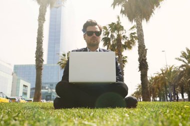 Güneşli gün laptop yeşil çimenlerin üzerinde çalışan resmi giysili genç beyaz iş adamı. Arka plan bulanık.