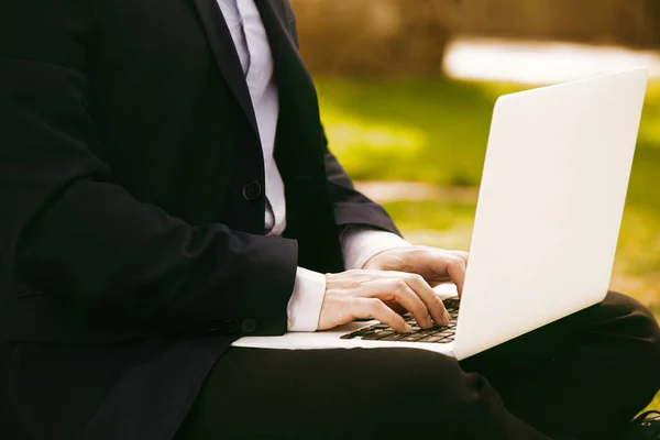 Närbild bild av ung affärsman i formalwear skriver på laptop tangentbord medan du sitter vid stadens gröna gräs utomhus. Suddig bakgrund. — Stockfoto