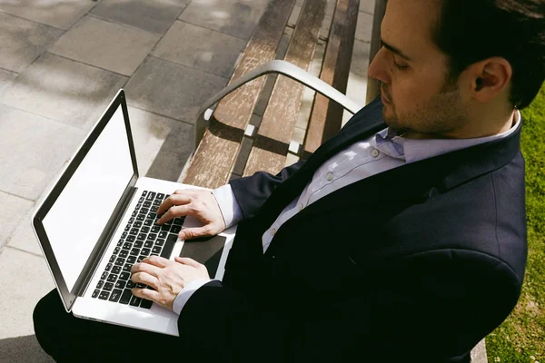 Närbild bild av ung affärsman i formalwear maskinskrivning på laptop tangentbord medan du sitter på bänken utomhus. Suddig bakgrund. — Stockfoto