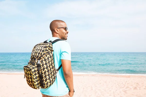 Retrato de un atractivo joven hipster afroamericano con camiseta y gafas de sol caminando en el día de verano en la playa con mochila . —  Fotos de Stock