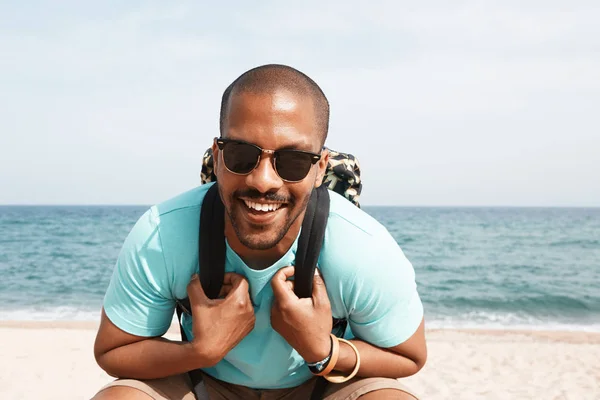 Feliz joven afroamericano hipster con camiseta y gafas de sol caminando en el día de verano en la playa con mochila . —  Fotos de Stock
