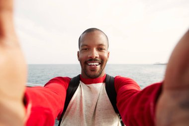 Mutlu yüz açık havada City ile iki el selfie alarak gülümseyen genç Afro-Amerikan hipster yakışıklı. Mavi arka plan bulanık denizde.