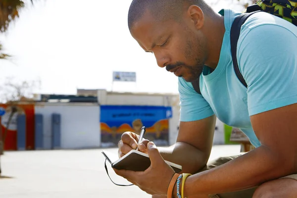 Atractivo hombre hipster afroamericano pensativo con mochila y tomando notas en copybook en el parque de la ciudad. Estudiante preparándose para la lección en la universidad . —  Fotos de Stock