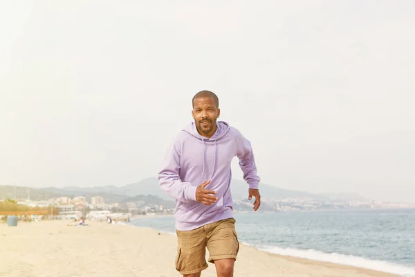 Concepto deportivo activo de verano. Atractivo feliz joven afroamericano hipster hombre en el deporte sudadera con capucha corriendo en la playa . —  Fotos de Stock