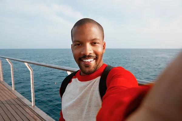 Guapo y sonriente joven hipster afroamericano tomando selfie con cara feliz en la playa. Mar azul sobre el fondo borroso . —  Fotos de Stock