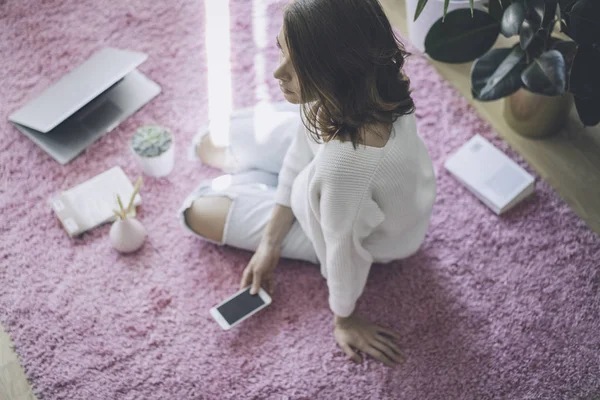 Mulher bonita sentada no tapete rosa no chão na sala de estar moderna e de cor clara com gadgets eletrônicos . — Fotografia de Stock