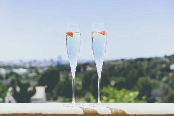 Two champagne glasses with strawberry on sunny terrace outdoor patio overlooking green trees at summer day outside of the city.