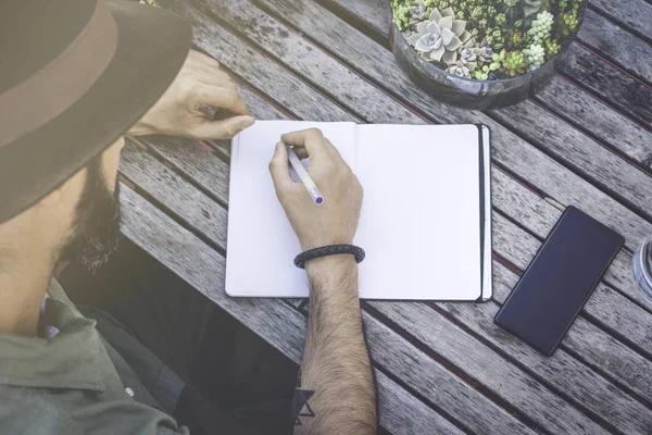 Hipster studentowi dokonywanie notatek, pisanie cele w Notatniku, siedząc na tarasie kawiarni. Skoncentrowane człowiek pisania wiadomości na biurko z drewna. Widok z góry. Efekt filmu — Zdjęcie stockowe