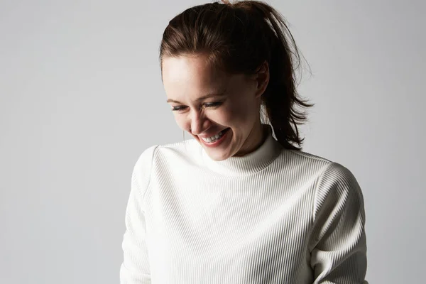 Fashion photo of a beautiful smiling brunette woman in a white roll-neck sweater posing over gray empty background. Fashionable photo shot. — Stock Photo, Image