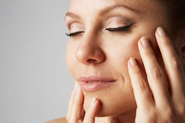 Beautiful handsome woman with clean skin, natural make-up, and closed eyes touching hands her face on gray background. Close up portrait.Cropped. — Stock Photo, Image