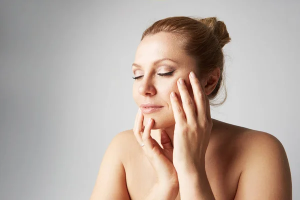 Beautiful mature woman with clean skin, natural make-up, and closed eyes touching hands her face on gray background. Close up portrait. Medical and cosmetic facial skin care concept. — Stock Photo, Image