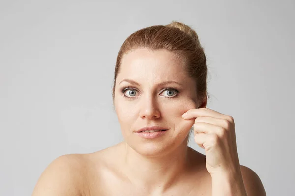 Mulher jovem com pele limpa, maquiagem natural e olhos bonitos toca os dedos de seu rosto no fundo cinza. Conceito de cuidados médicos e cosméticos com a pele facial . — Fotografia de Stock