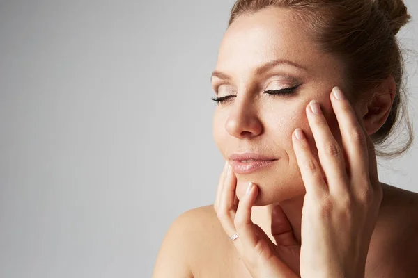 स्वच्छ त्वचेसह सुंदर हस्तमैथुन स्त्री, नैसर्गिक मेक-अप आणि बंद डोळे राखाडी पार्श्वभूमीवर तिचा चेहरा स्पर्श करतात. चित्र बंद करा. वैद्यकीय आणि सौंदर्यप्रसाधन चेहर्याचा त्वचा काळजी संकल्पना . स्टॉक इमेज
