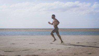 Genç adam deniz kıyısında güneş doğarken koşu sakallı. Parlak güneş ışığı ile plajda koşan sağlıklı genç sakallı adam portresi yan