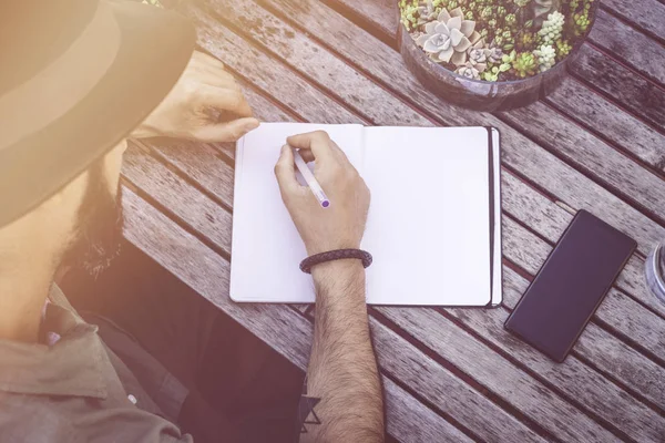 Hipster studentowi dokonywanie notatek, pisanie cele w Notatniku, siedząc na tarasie kawiarni. Skoncentrowane człowiek pisania wiadomości na biurko z drewna. Widok z góry. Makieta flary — Zdjęcie stockowe