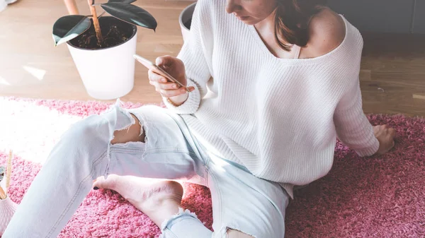 Menina bonita sentada no tapete rosa no chão na moderna sala de estar de cor clara e mão de digitação no telefone móvel . — Fotografia de Stock