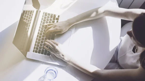 Werkende momenten op naaiatelier. Jonge vrouw te typen op laptopcomputer zittend aan de houten tafel in licht gekleurd woonkamer van moderne huis — Stockfoto