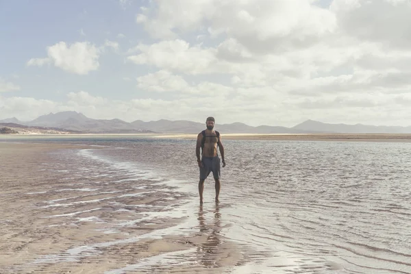 Young muscular bearded man standing on the sea shore at sunrise. Side portrait of healthy young bearded man running at the beach with bright sunlight