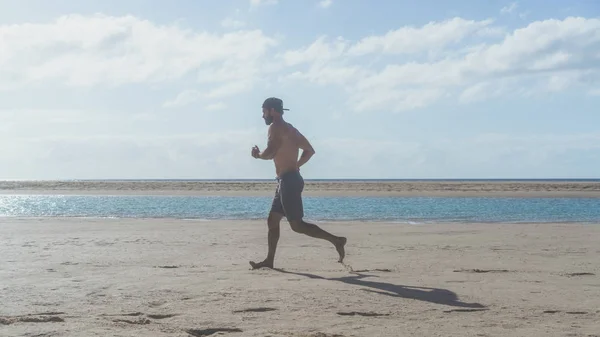 Feliz hombre barbudo corriendo en la orilla del mar al amanecer. Retrato lateral de un joven barbudo sano corriendo por la playa con luz solar brillante —  Fotos de Stock