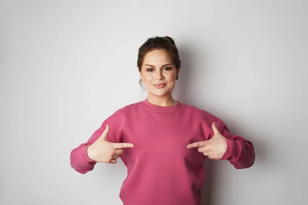 Beautiful smiling hipster girl wearing pink hoody and pointing finger with space for your logo or design. Mock-up of pink hoody on white empty wall in the background. Copy paste text space. — Stock Photo, Image