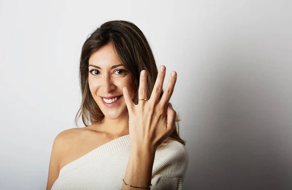 Happy european girl with long dark hair in stylish clothes smiling and holding hand on hip while showing trendy wedding ring over empty white wall background.Copy paste text message space. — Stock Photo, Image