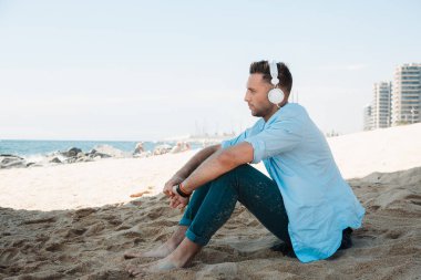 Genç hipster adam mavi tişört ve kot kulaklık bir smartphone cep telefonu ile müzik dinlemek ve denizde seyir sahil kumu üzerinde oturuyor. Dijital müzik salonu ve rahatlatıcı kavramı.
