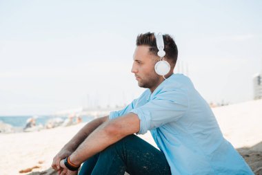 Genç hipster adam mavi tişört ve kot kulaklık bir smartphone cep telefonu ile müzik dinlemek ve denizde seyir sahil kumu üzerinde oturuyor. Dijital müzik salonu ve rahatlatıcı kavramı. Kırpılmış.