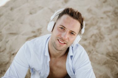 Genç hipster adam beyaz bir gömlek ve bir akıllı telefon kulaklıktan müzik kot portresi ve sahilde oturuyor. Dijital müzik salonu ve rahatlatıcı kavramı. Portre.