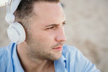 Genç hipster adam mavi tişört ve kot bir smartphone kulaklıktan müzik portresi ve sahilde oturuyor. Dijital müzik salonu ve rahatlatıcı kavramı. Portre.