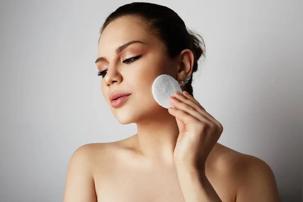 Hermosa chica con grandes ojos marrones y cejas oscuras refrescante cara de piel con almohadillas de algodón sobre fondo de estudio grises. Modelo con maquillaje desnudo ligero. . Imagen de archivo